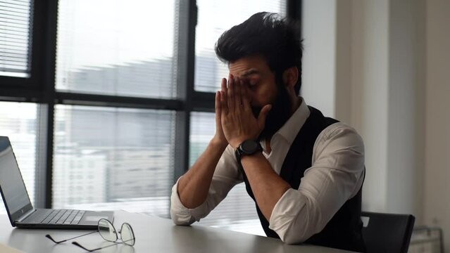 Exhausted overworked business man take off eyeglasses and massaging dry itchy irritated eyes working at office desk with laptop near window. Businessman rubbing massaging neck suffering from pain.