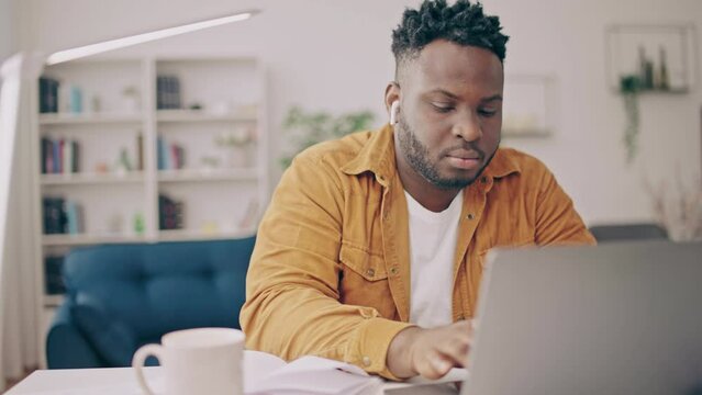 Concentrated black man typing on laptop and taking notes in notepad, freelance