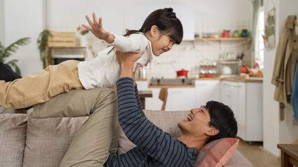 happy Asian dad having fun lifting up daughter on straight legs while playing airplane on living room couch at home. father doing yoga with child concept