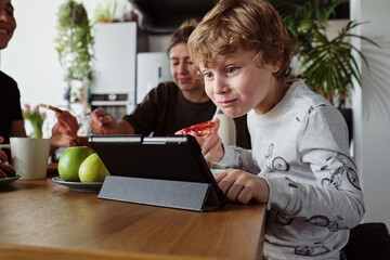 Little boy watching cartoons on tablet pc during breakfast with family at table