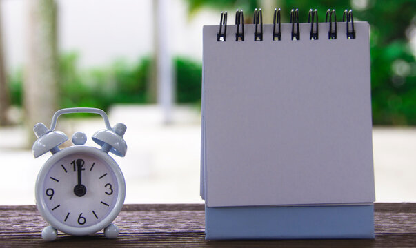 Alarm clock pointing at 12 o'clock with blank notepad. Blurred park background. Copy space