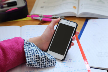 Ein Mobiltelefon in der Hand mit Stoffmaske am Handgelenk. Im Hintergrund sind Lehrbuch, Hefte,...
