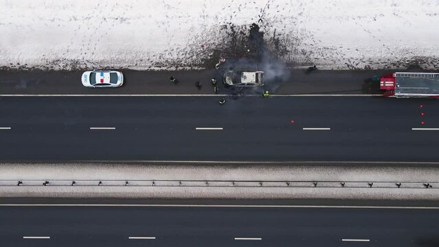 Accident - burned out car on the edge of the motorway in winter, aerial view. Near the fire and police cars. Help and rescue concept, 911 service. White snow on the side of the road. UHD 4K