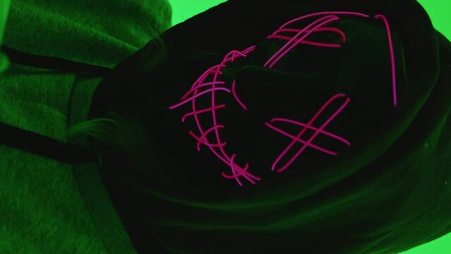 Vertical video, Jason style mask, cryptic horror with pink fluorescent neon lights and green chroma key background. Costume party wearing a black hood.