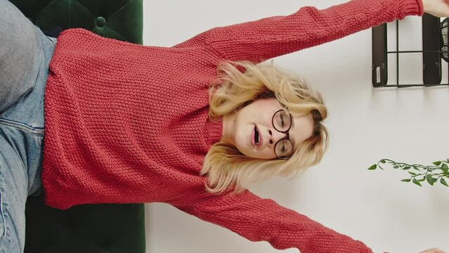 Vertical video, Young blonde woman wearing a red sweater and glasses stretches, takes a deep breath and yawns sitting on a green sofa, in the living room of a house with hanging plants.