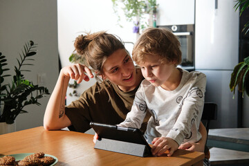 Young mother talking to her son while he playing digital tablet at table in kitchen