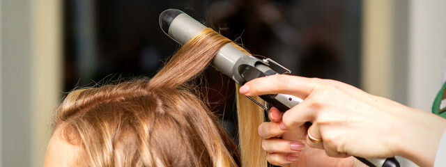 Fototapeta premium Hairdresser makes curls with a curling iron for the young woman with long brown hair in a beauty salon