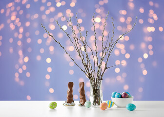 easter, holidays and object concept - pussy willow branches, colored eggs and chocolate bunnies on table over festive lights on violet background © Syda Productions