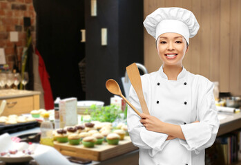 cooking, culinary and people concept - happy smiling female chef in toque with wooden spoon over restaurant kitchen background