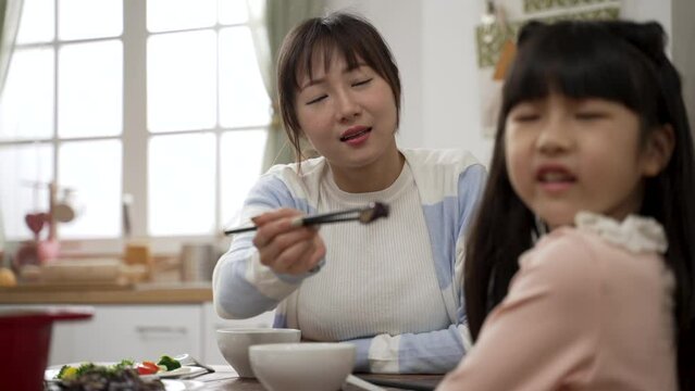 selective focus of frustrated Asian mother trying to encourage daughter to eat vegetable in dining room at home. the girl turns head and refuses her worried mom