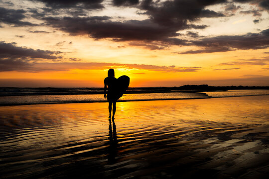 Surfer girl on the beach at sunset. Surf silhouette of woman walking with surfboard at golden hour beautiful colors