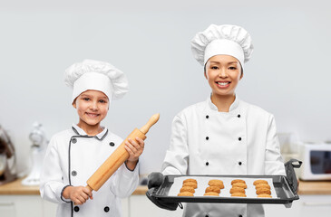 cooking, culinary and bakery concept - happy smiling female chef or baker in toque holding baking tray with oatmeal cookies and little girl with rolling pin over kitchen background