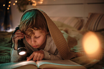 Child lying under blanket on bed using flashlight to read a fairy tale before sleep - obrazy, fototapety, plakaty