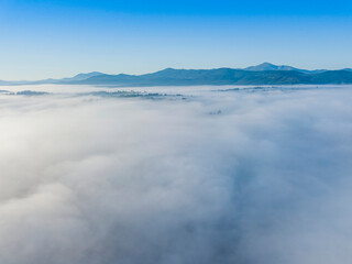 Flight over fog in Ukrainian Carpathians in summer. Mountains on the horizon. A thick layer of fog covers the mountains with a continuous carpet. Aerial drone view.