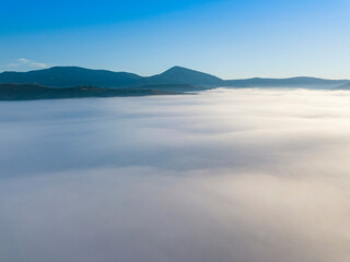 Flight over fog in Ukrainian Carpathians in summer. Mountains on the horizon. A thick layer of fog covers the mountains with a continuous carpet. Aerial drone view.