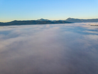 Obraz premium Flight over fog in Ukrainian Carpathians in summer. Mountains on the horizon. A thick layer of fog covers the mountains with a continuous carpet. Aerial drone view.
