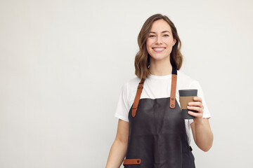 Portrait of beautiful young barista woman looking in camera and smiling, while holding a to-go...