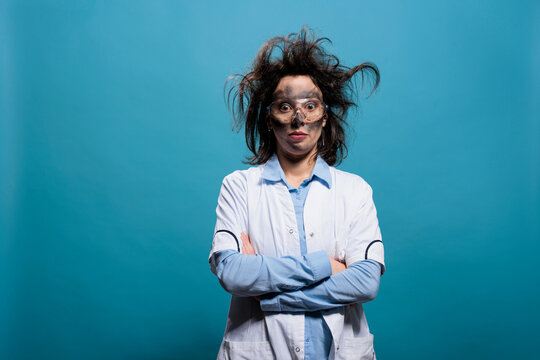 Humorous mad scientist with erratic hair and filthy face standing with crossed arms. Hilarious looking crazy chemist with messy hairstyle and dirty face standing on blue background looking at camera.