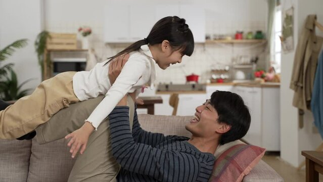 Side view happy Asian father lying on sofa and holding up daughter on his legs at home. The girl open arms and flying in air like plane