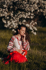 Walks in nature in the countryside. Close-up portrait of a young woman in a blooming garden. A girl in a national costume, a bandage with a flower on her head, an embroidered shirt and a red skirt.