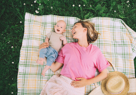 Smiling mom and baby lying on blanket on green grass at summer. Family relaxing and having picnic outdoors. Beautiful mother with her baby on nature. Concept of motherhood, human happiness, eco life. - Powered by Adobe