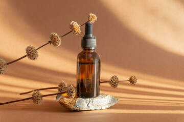 Mockup of amber glass bottle with facial serum on granite stone stand. Container with dropper lid surrounded by dry twigs. Essential oil for care of women's skin on brown background