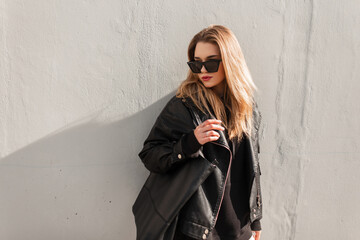 Fashionable pretty woman hipster with stylish sunglasses in black leather jacket with fashion handbag stands and poses near a gray wall in spring sunny day. Fashion, style and beauty