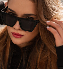Fototapeta premium Stylish portrait of a beautiful fashionable woman with trendy sunglasses in a black trendy vintage leather jacket and hoodie looking at the camera