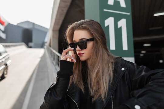 Fashion beautiful woman with trendy sunglasses in stylish rock leather jacket with black hoodie walks in the city near a parking lot