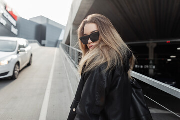 Stylish beautiful girl with cool glasses in a fashionable leather jacket and hoodie with a bag...