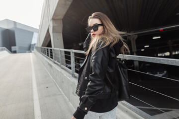 Fototapeta na wymiar Fashionable young girl with cool fashion sunglasses in trendy black leather clothes with leather rock jacket and bag walks in the city. Urban female spring style outfit