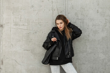 Fashion young beauty woman hipster model with rock leather jacket, hoodie and fashion leather bag stands near a concrete gray wall. Female urban style, fashion and beauty