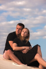 family relationship, romantic date. couple in black summer clothing  are sitting in hugs on a sand career and looking apart in desert at sunset on a blue sky background . lifestyle concept, free space