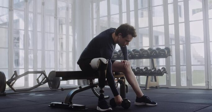 Caucasian male with prosthetic leg putting earphones in his ears before workout. Disabled athlete working out in modern-style gym.