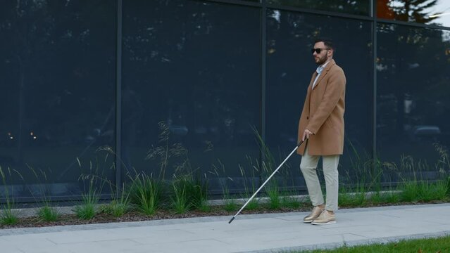 Caucasian blind man in stylish outfit using cane for safety walk on city street. Handsome male person checking direction with stick outdoors.