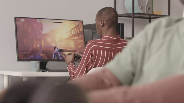 Rear view medium slowmo of African American man playing fps game on PC with controller while his colleague typing program code developing video games in blurred foreground
