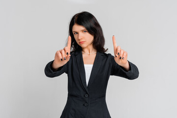 Please, do not, stop. Young serious dark-haired girl in black blazer, who is looking in the camera and pointing fingers up, standing over grey background