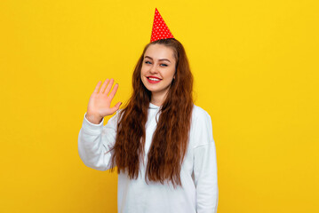 Friendly woman in celebratory cap, waving hand in hello, hi gesture, informal greeting of guests, smiling cheerful. Colorful studio portrait with yellow background. Holiday and party concept © Red Cristal