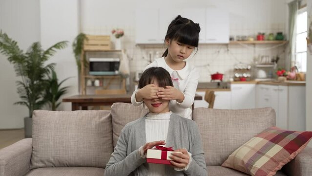 sweet Asian young daughter covering mom&rsquo;s eyes and giving her a surprise present in living room on mother&rsquo;s day. she hugs her happy mother from behind with smile