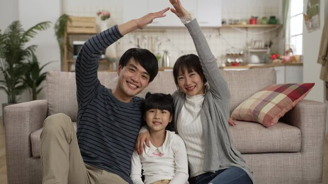 slow motion portrait of asian father and mother smiling at camera and making roof figure with hands over their heads while sitting close with their daughter at home