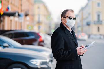 Serious businessman poses outdoor near transport at street, holds modern cellular and newspaper, dressed in black coat, wears protective mask from coronavirus.