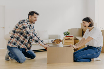 Fototapeta na wymiar Funny woman and man have fun during relocation, pose on floor with dog in cardboard box, just moved into apartment