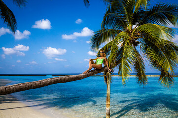 Beautiful woman in a bikini on a palm tree on the paradise beach of Maldives. Sexy model posing in a bikini on the beach. Luxury travels. Holidays on a paradise island. Maldives landscape.