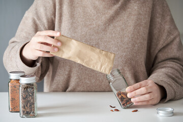 Unrecognizable woman refilling spice jar with dry cayenne pepper from a paper bag buyed at package free grocery store.