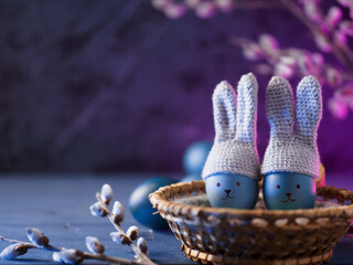 blue eggs in crocheted hats with bunny ears and painted muzzles in a wicker basket, willow branches. neon background with copy space