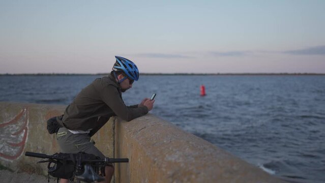 Theme is cycling in seaside. Male cyclist stopped to enjoy the sea view and see the road on the map on his smartphone. A male with a bicycle on the embankment is resting and using a smart phone.