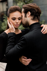 Portrait of fashion young adult couple on the photoshoot, looking directly at the camera. Handsome beard man in stylish suit hugging gorgeous woman in black dress, posing for the photo