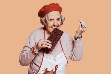 Funny portrait of cheerful overweight elderly cook grandmother biting into a chocolate bar, looking at camera at studio over beige background. Copy space.