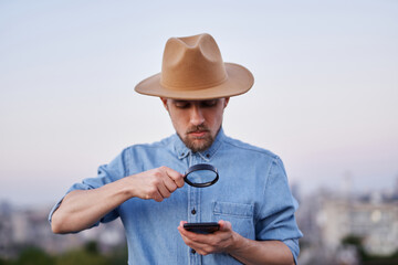 Man wearing jeans shirt and hat looking with loupe at mobile phone as detective or investigator concept. Cute bearded male looking with magnifying glass at smartphone. High quality image