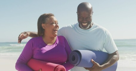 Portrait of happy senior african american couple holding yoga mats on sunny beach
